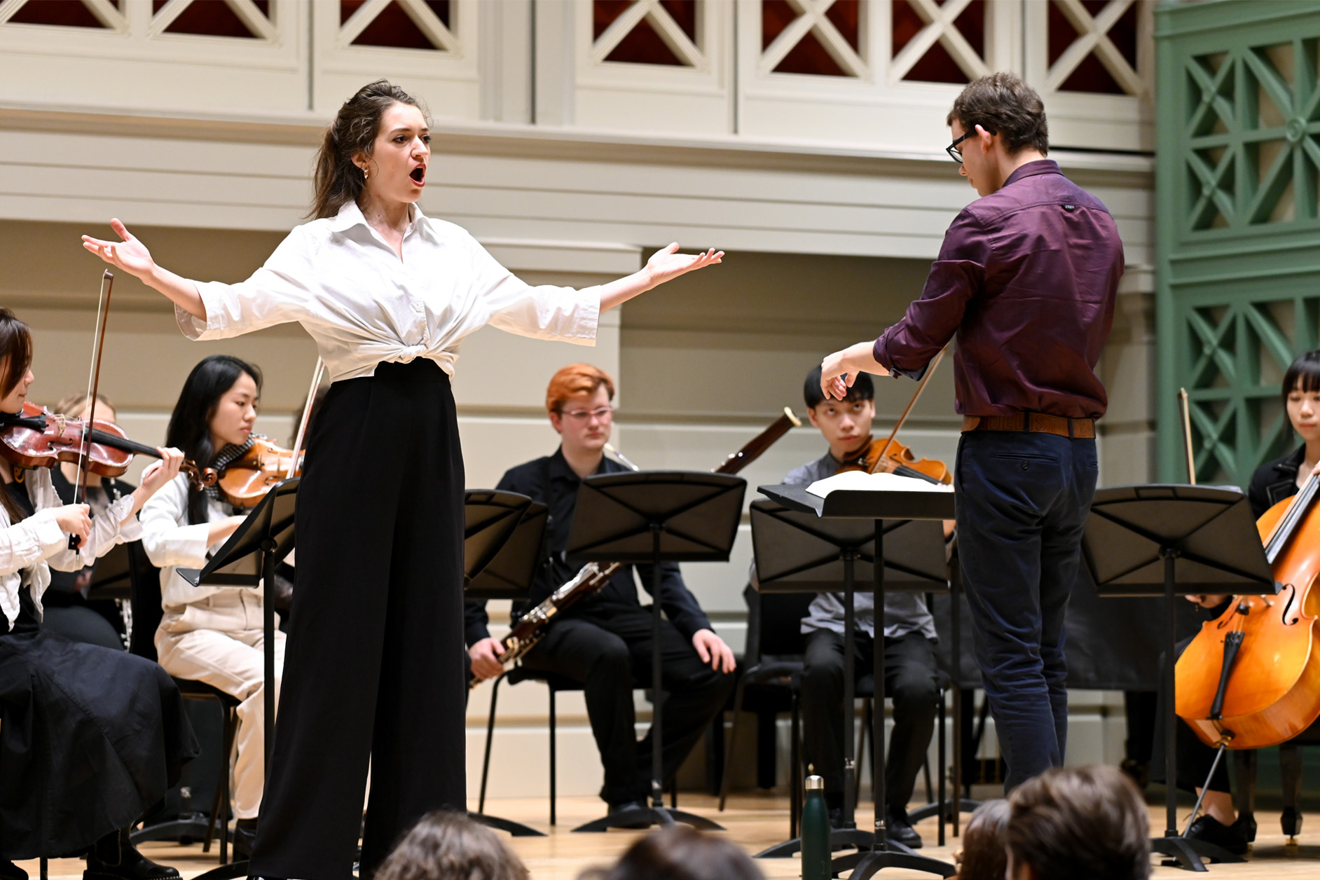 A female singer, wearing a white shirt and black trousers, performing on stage with an ensemble behind her, standing next to the conductor